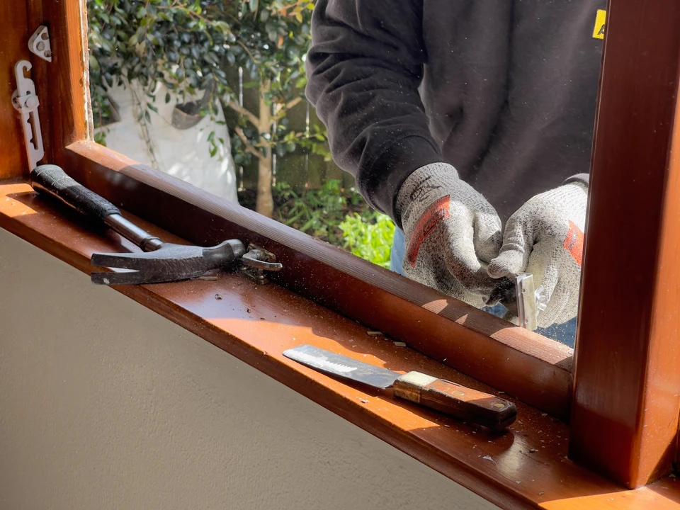 Artisan menuisier en atelier appliquant une finition à la brosse sur le cadre d'une fenêtre en bois massif posée horizontalement sur un établi de travail, concentré sur le détail du travail, avec des outils de menuiserie et des pièces de bois en arrière-plan.